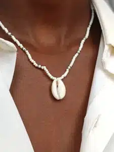 Gros plan sur un collier en perles porté autour du cou. Le collier est composé de petites perles blanches et dorées, et au centre pend un coquillage de type cauri. La personne porte une chemise blanche entrouverte.