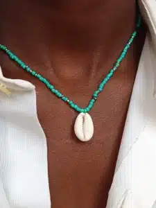 Gros plan sur un collier en perles porté autour du cou. Le collier est composé de petites perles dorées et turquoises , et au centre pend un coquillage de type cauri. La personne porte une chemise blanche entrouverte.