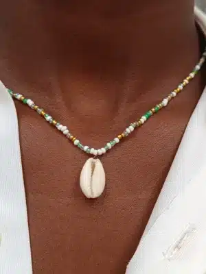 Gros plan sur un collier en perles porté autour du cou. Le collier est composé de petites perles blanches, dorées et vertes, et au centre pend un coquillage de type cauri. La personne porte une chemise blanche entrouverte.