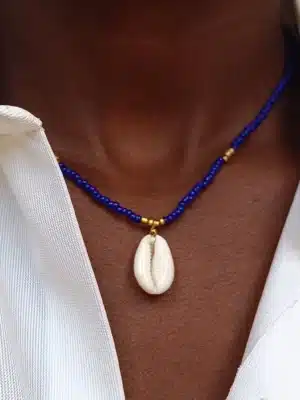 Gros plan sur un collier en perles porté autour du cou. Le collier est composé de petites perles bleu et dorées , et au centre pend un coquillage de type cauri. La personne porte une chemise blanche entrouverte.