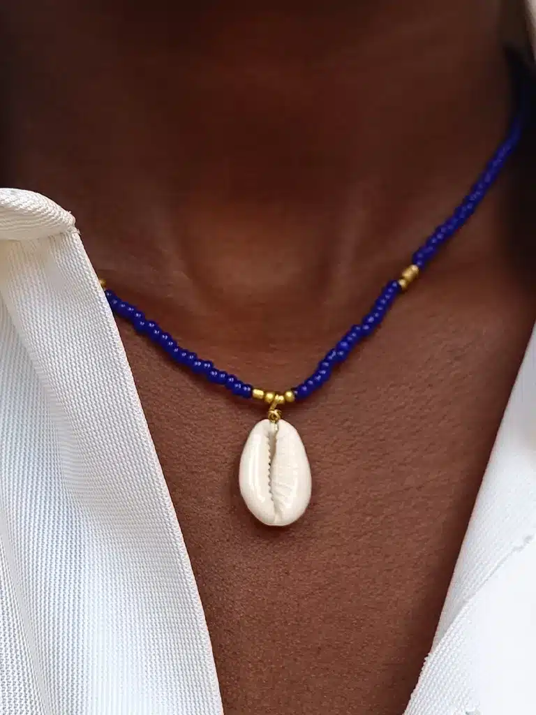 Gros plan sur un collier en perles porté autour du cou. Le collier est composé de petites perles bleu et dorées , et au centre pend un coquillage de type cauri. La personne porte une chemise blanche entrouverte.