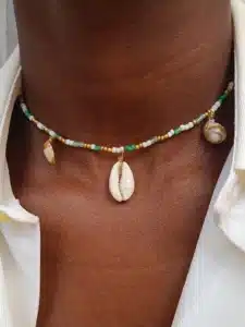 Gros plan sur un collier en perles porté autour du cou. Le collier est composé de petites perles blanches, vertes et dorées, ainsi que de quelques petites perles naturelles. Un coquillage de type cauri pend au centre comme pendentif. La personne porte une chemise blanche légèrement ouverte.