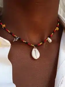 Gros plan sur un collier en perles porté autour du cou. Le collier est composé de petites perles rouges, noires et dorées, ainsi que de quelques petites perles naturelles. Un coquillage de type cauri pend au centre comme pendentif. La personne porte une chemise blanche légèrement ouverte.