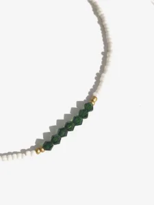 Colliers délicats faits de petites perles blanches, ornés au centre de perles facettées et vertes, séparées par de petites perles dorées, posés sur un fond blanc avec une touche de paille séchée en arrière-plan.