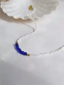 Colliers délicats faits de petites perles blanches, ornés au centre de perles facettées bleues, séparées par de petites perles dorées, posés sur un fond blanc avec une touche de paille séchée en arrière-plan.