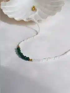 Colliers délicats faits de petites perles blanches, ornés au centre de perles facettées vertes, séparées par de petites perles dorées, posés sur un fond blanc avec une touche de paille séchée en arrière-plan.