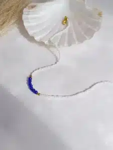 Colliers délicats faits de petites perles blanches, ornés au centre de perles facettées bleues , séparées par de petites perles dorées, posés sur un fond blanc avec une touche de paille séchée en arrière-plan.