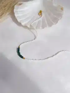 Colliers délicats faits de petites perles blanches, ornés au centre de perles facettées bleues, séparées par de petites perles dorées, posés sur un fond blanc avec une touche de paille séchée en arrière-plan.