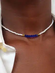 Gros plan sur un collier minimaliste africain moderne en perles porté autour du cou. Le collier est composé de petites perles blanches rehaussées par des petites perles dorées. , Au centre une rangé de perles en cristal bleu . La personne porte une chemise blanche entrouverte.