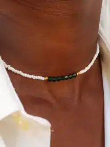 Gros plan sur un collier minimaliste africain moderne en perles porté autour du cou. Le collier est composé de petites perles blanches rehaussées par des petites perles dorées. , Au centre une rangé de perles en cristal vert . La personne porte une chemise blanche entrouverte.